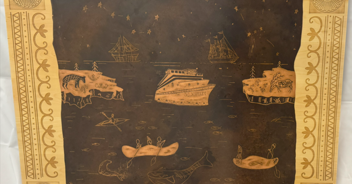 Plaque en bois gravée à bord du navire Ala'suinu, illustrant des œuvres d'art de style mi'kmaq représentant des navires, des canots, la faune et les étoiles, accompagnée d'une inscription expliquant le nom du navire.